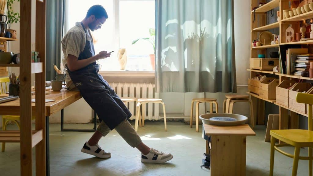 A man in an apron leans on a table while looking at his phone in a workshop.