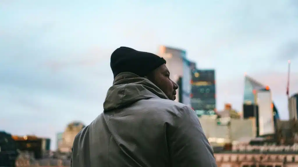 A man in a beanie and jacket looks out at city buildings.