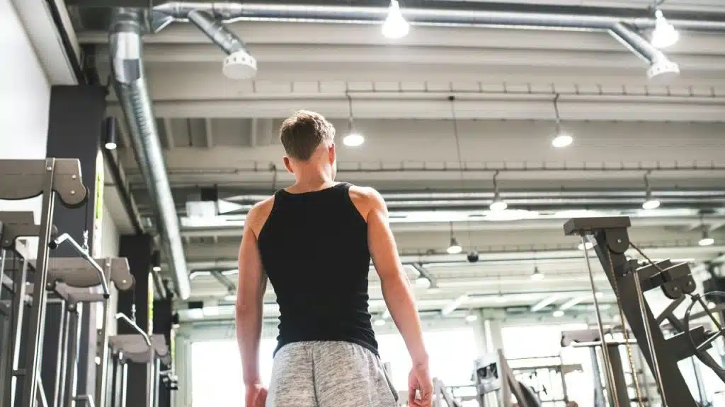 A man stands in a gym wearing a black tank top.