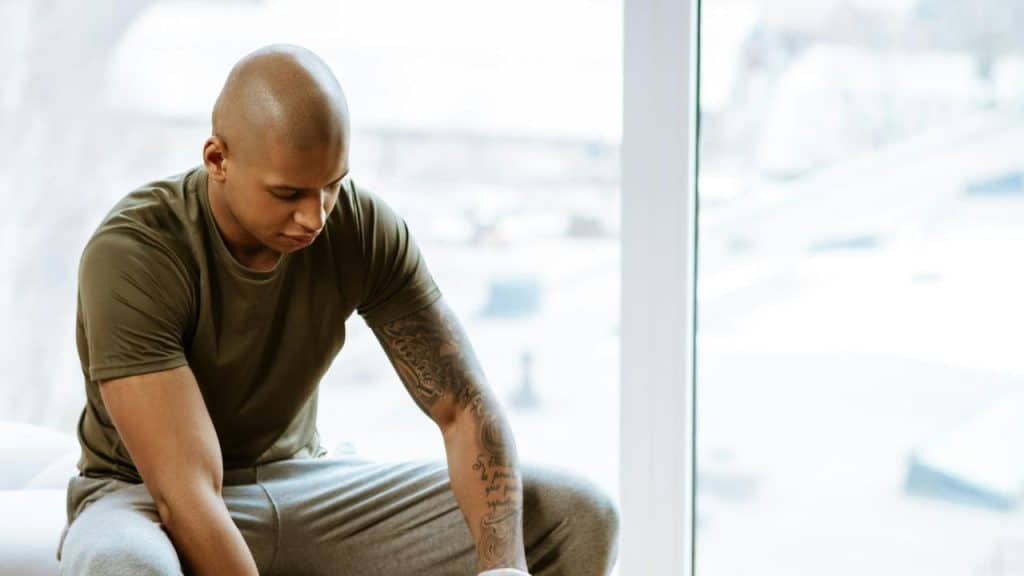 A man with a tattoo sits indoors looking down.