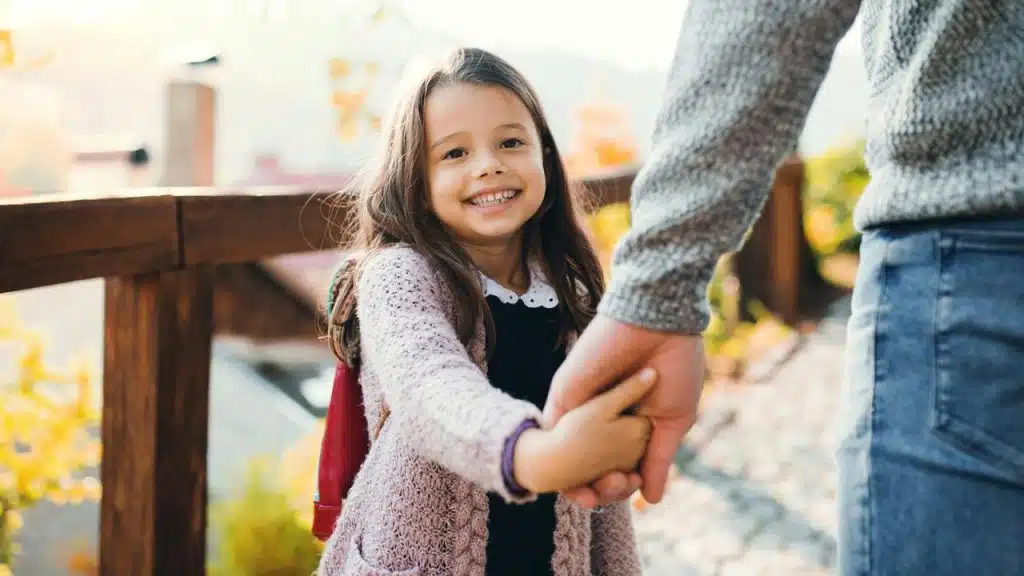 A smiling girl holds an adult’s hand outdoors.