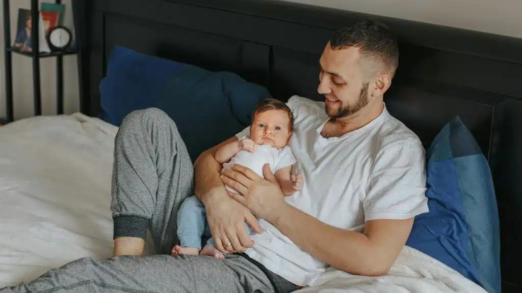 A father smiles while holding his baby on a bed.