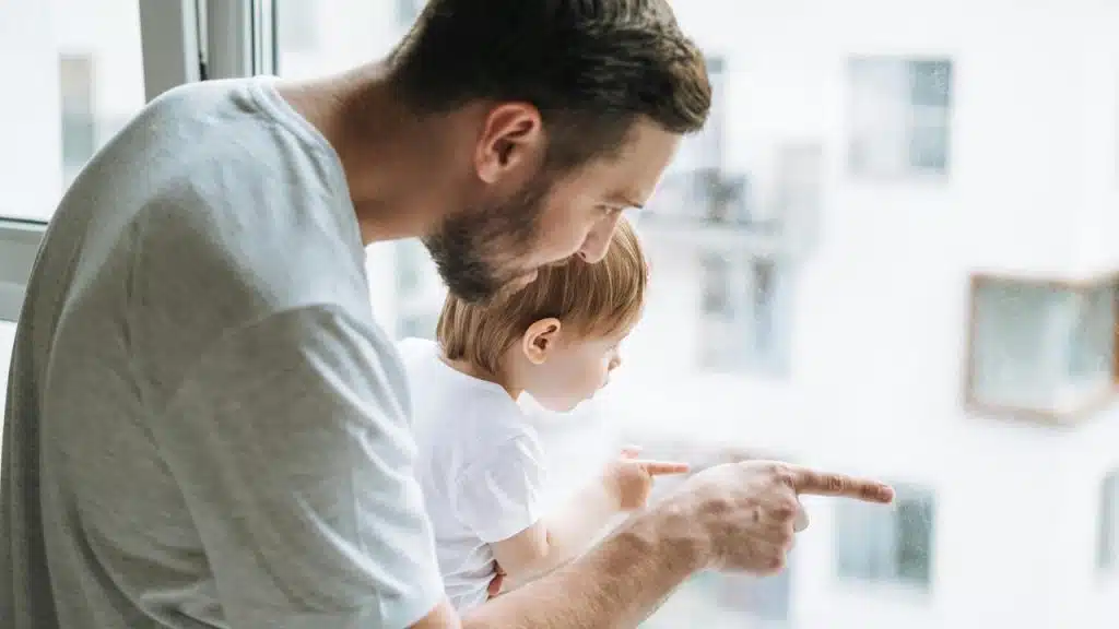 A father holds his baby while pointing out the window.