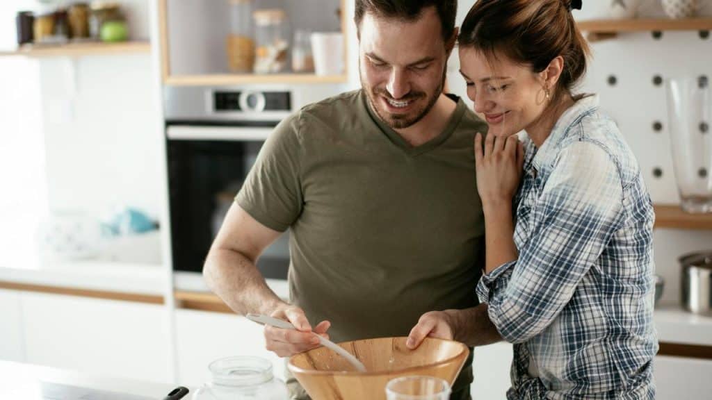 A couple smiling while preparing food together in a kitchen.
