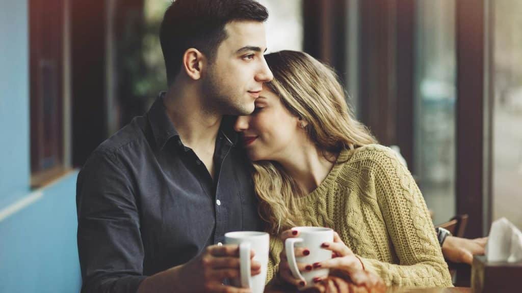 A couple sitting close together holding coffee mugs.