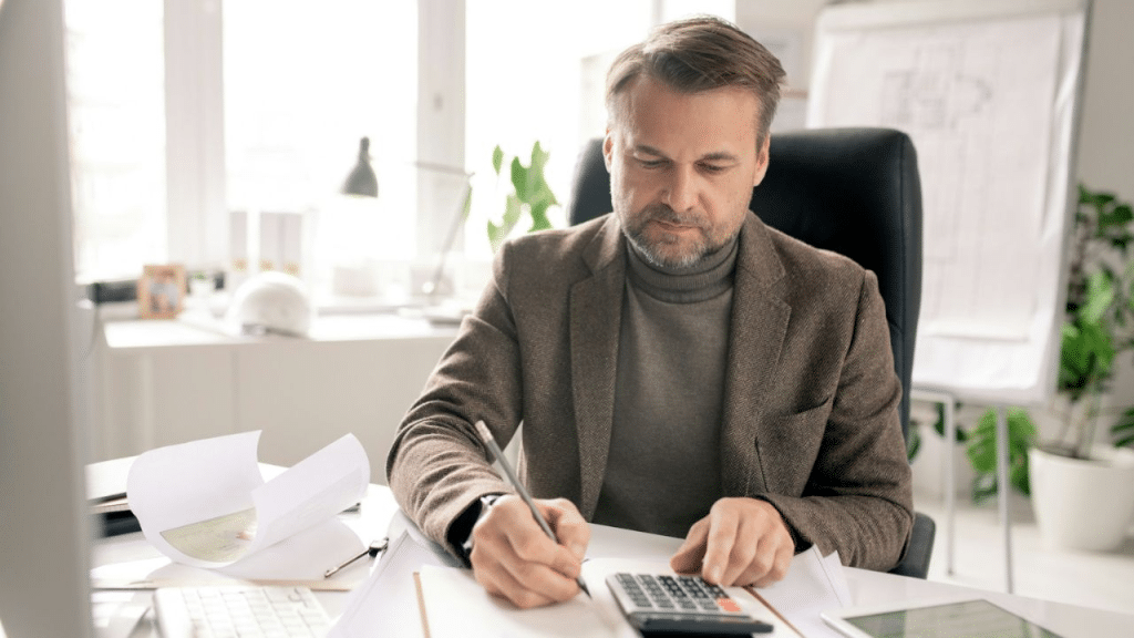 A serious, middle-aged man with a beard is working at his desk, using a calculator and writing.