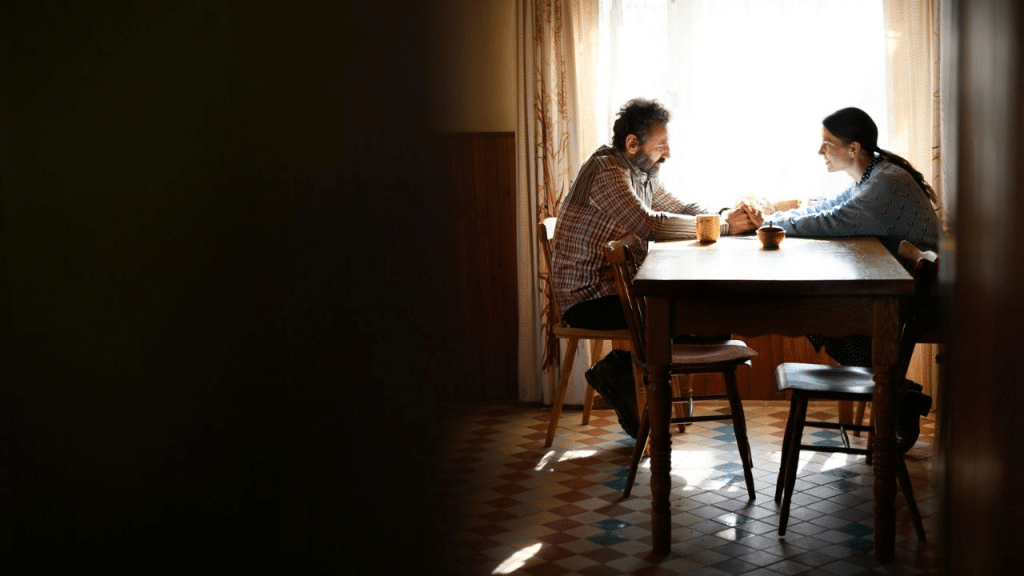 A middle-aged couple sits at a wooden table and holds hands, backlit by a large window.