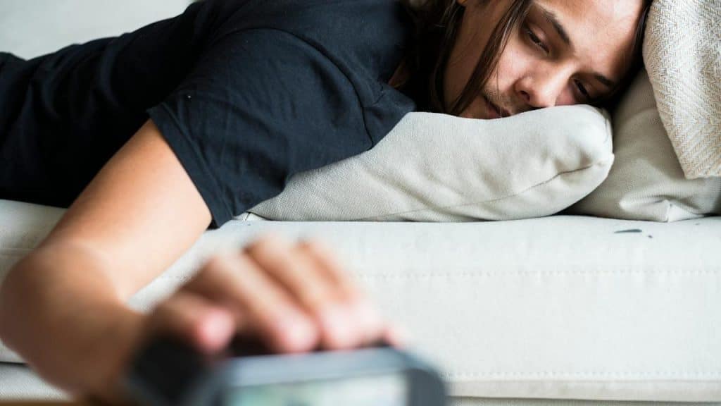 A man lying on a couch reaching for an alarm clock.