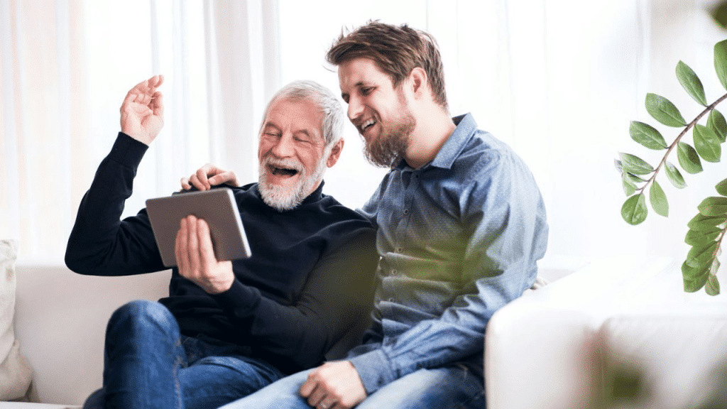 A smiling elderly man and a younger man with a beard are looking at a tablet together on a couch.