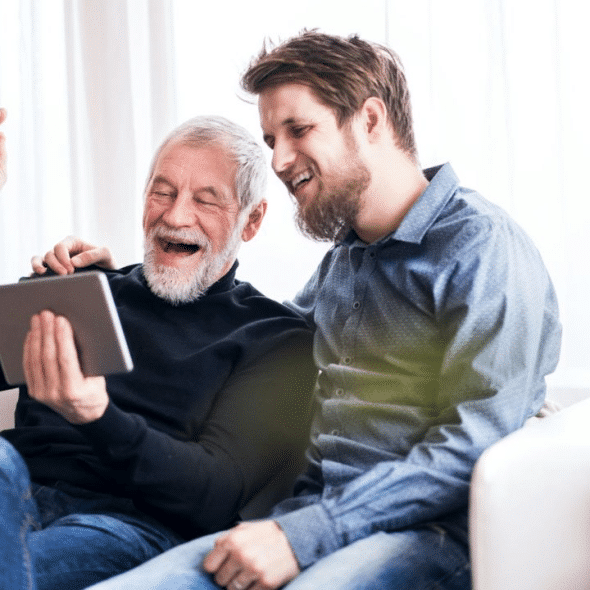 A smiling elderly man and a younger man with a beard are looking at a tablet together on a couch.