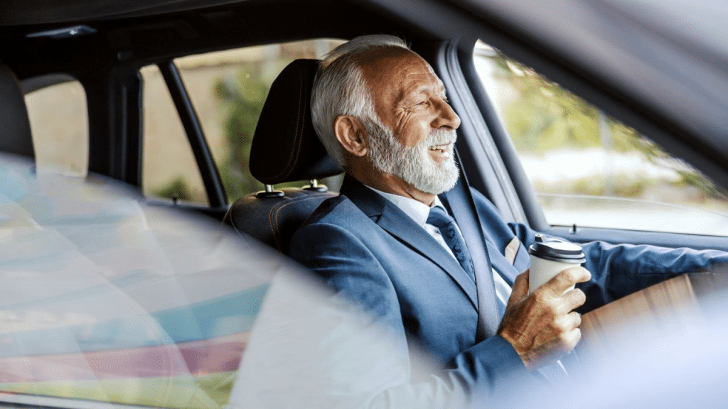 A cheerful elderly businessman with a white beard is driving a car and holding a cup of coffee.