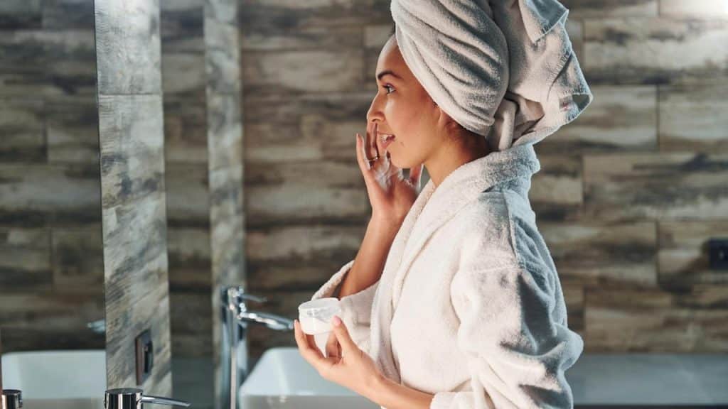 A woman in a bathrobe and towel applies skincare cream in front of a mirror.