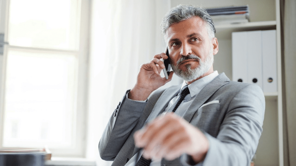 A serious, middle-aged man with gray hair and a beard is sitting at a desk and talking on a phone.