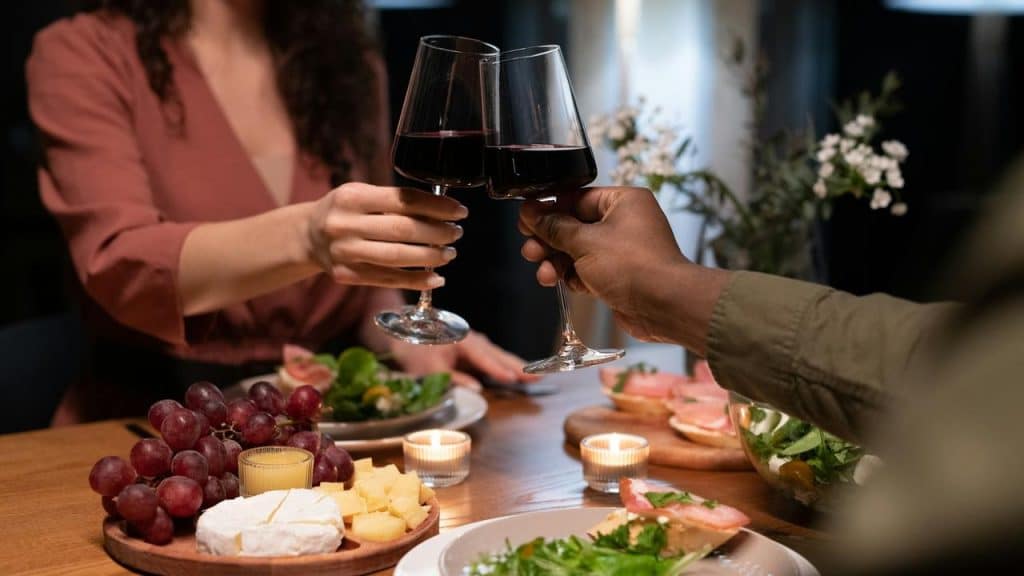 A couple clinking wine glasses over a candlelit dinner with cheese and grapes.