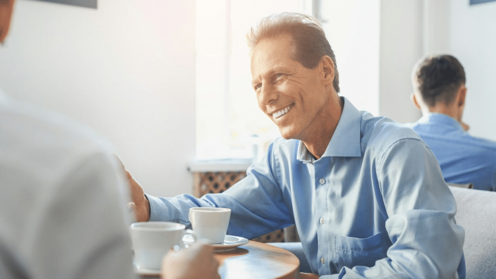 A handsome, smiling, middle-aged man in a blue shirt is having a conversation with someone while sitting at a coffee shop.