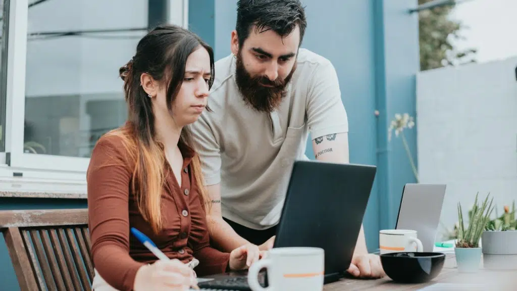 A man with a beard leans over a woman, looking at a laptop computer. She is writing in a notebook.