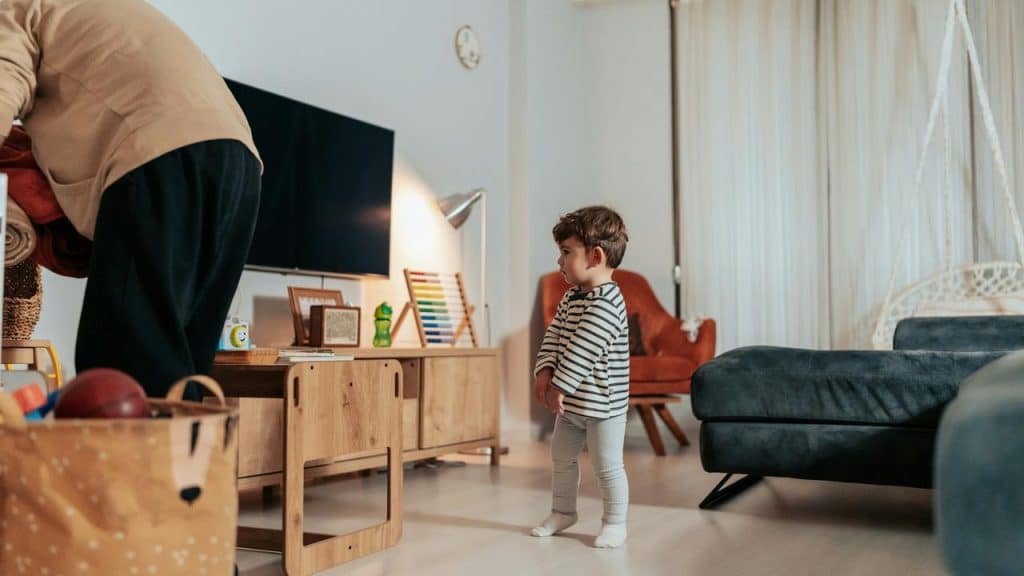 A toddler standing in a living room watching an adult tidy up.