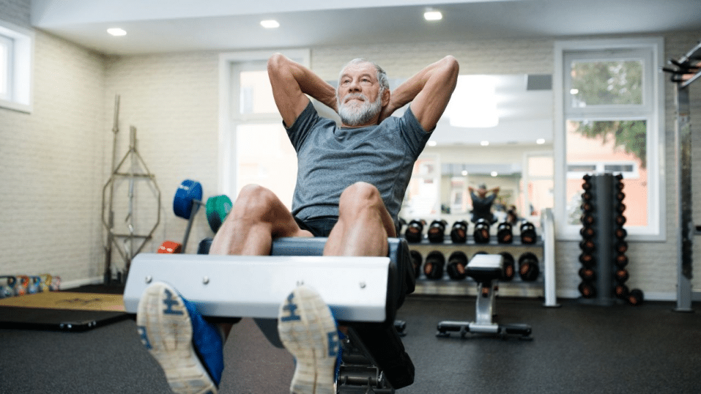 A smiling elderly man with a white beard does sit-ups on a workout bench.
