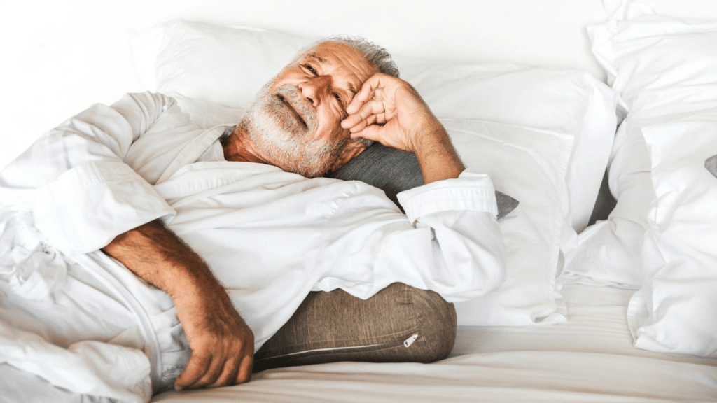 A content elderly man with a white beard lies on a white bed and smiles.