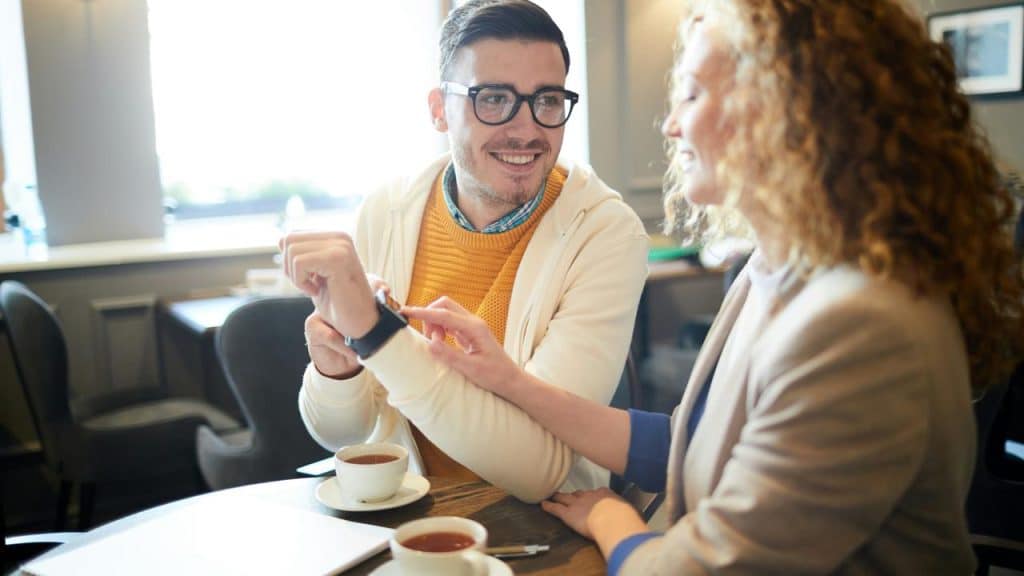 A man and woman sitting at a café table smiling while looking at a smartwatch.