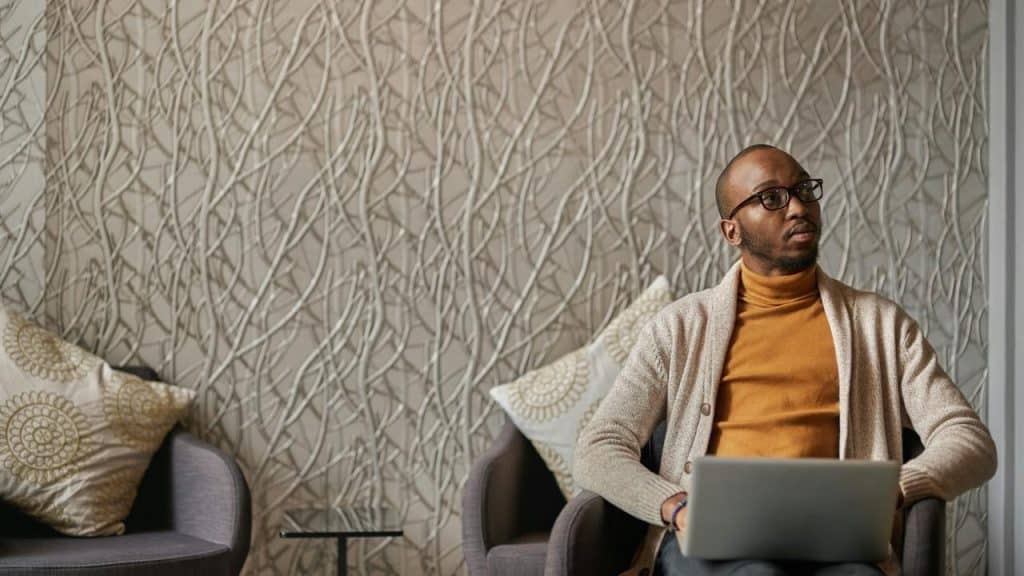 A man sitting with a laptop, looking to the side thoughtfully.