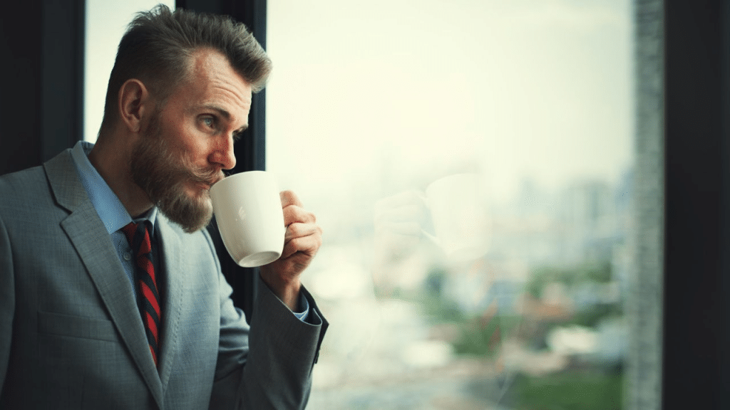 A bearded man in a gray suit and red tie sips from a white mug while looking out a window.