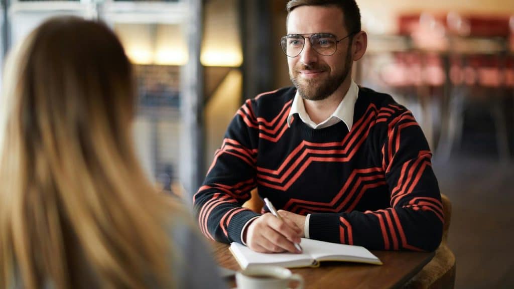 A man writing in a notebook while talking to a woman at a café.