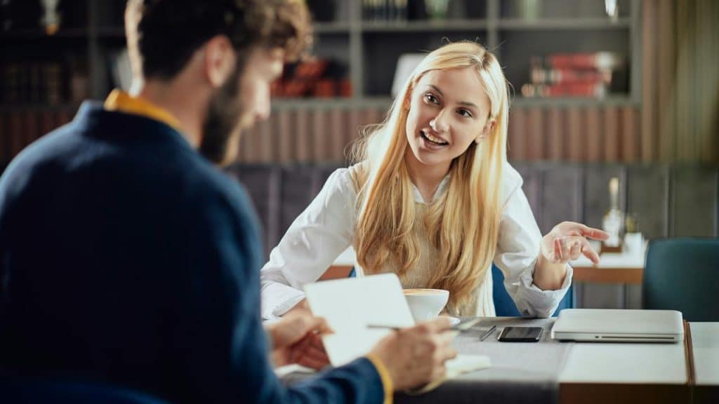 A woman smiling and talking to a man at a table with notebooks and coffee.