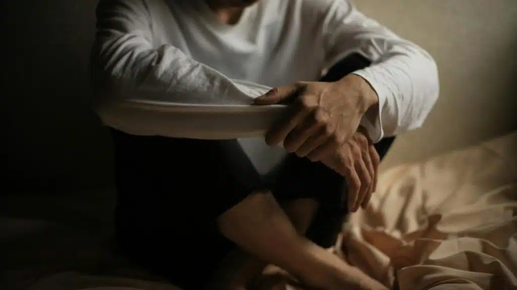 A person sits on a bed with arms resting on their knees, appearing thoughtful.