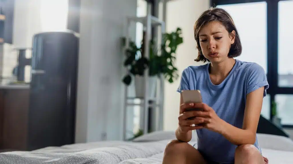 A woman sits on a bed looking at her phone with a worried expression.