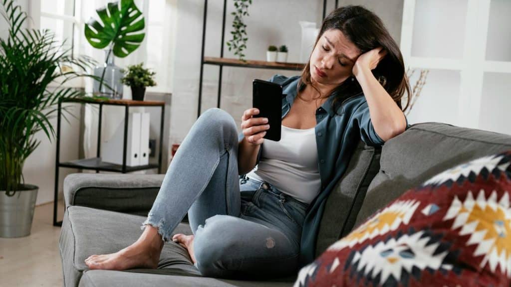 A woman sits on a couch looking at her phone with a stressed expression.