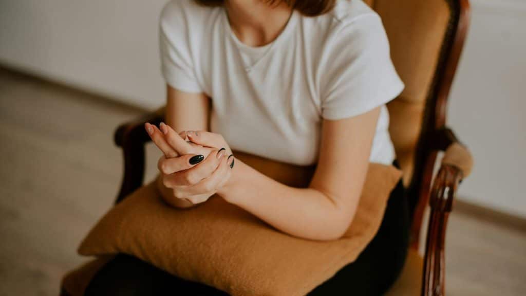 A woman sits on a chair holding her hands together with a pillow on her lap.