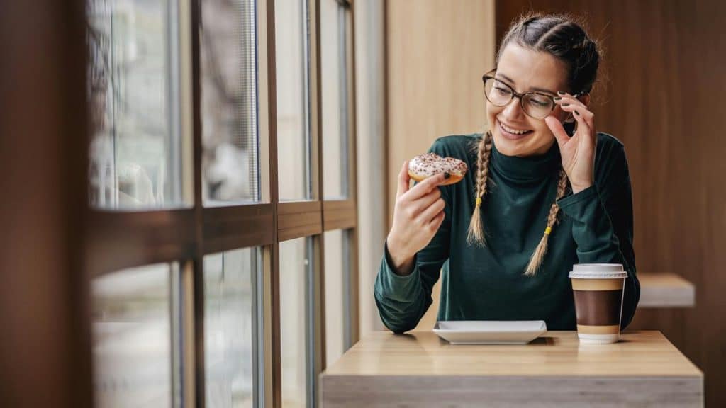 A woman with braids and glasses smiles while holding a donut and sitting with a coffee.