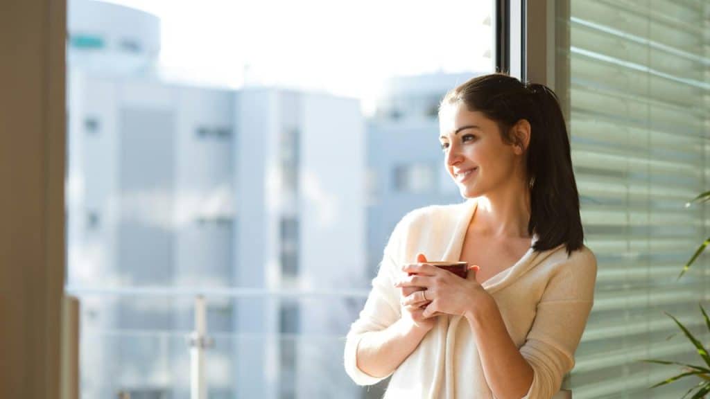 A woman stands by a window smiling while holding a cup.