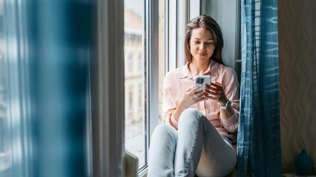 A woman with short hair smiles while holding a cup and sitting by a window.