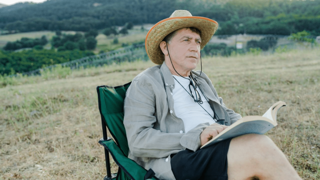 A middle-aged man in a straw hat and sunglasses is sitting on a portable chair outdoors, reading a book.