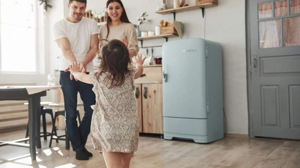 A little girl runs toward her smiling parents in a cozy kitchen.
