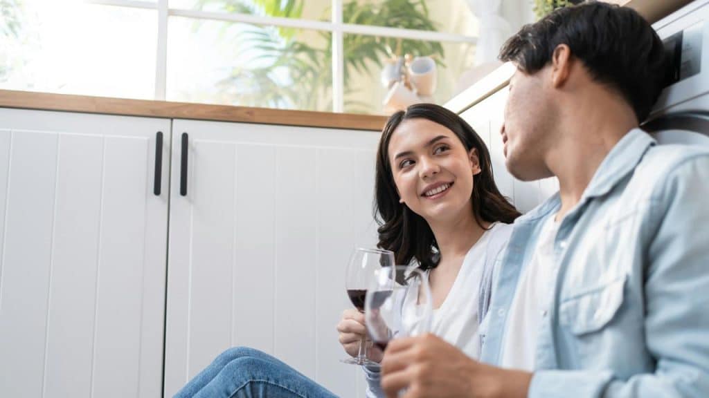 A couple sits on the floor smiling at each other while holding glasses of wine.