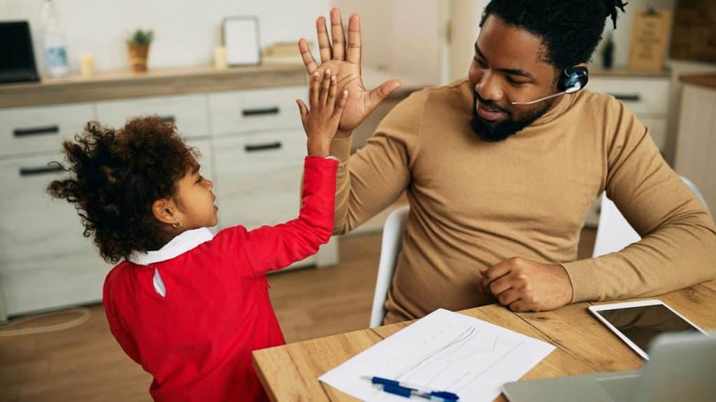 A father wearing a headset gives his daughter a high five while working at a desk.