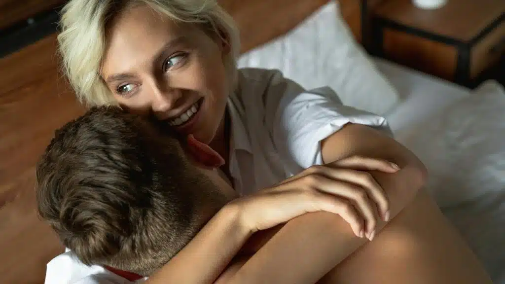 A woman smiles while hugging a man closely in bed.