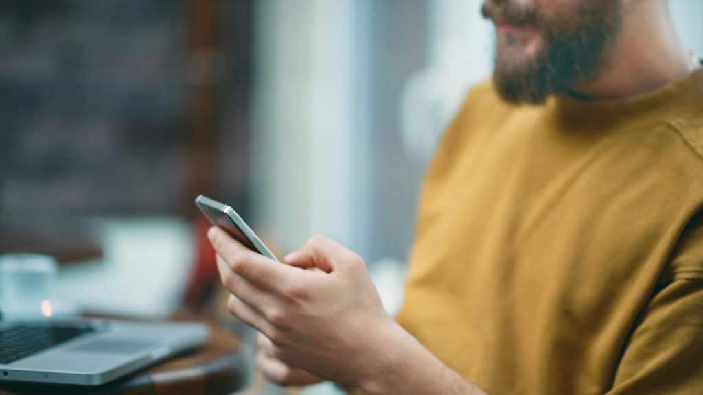 A man in a mustard sweater uses a smartphone while sitting at a table with a laptop.