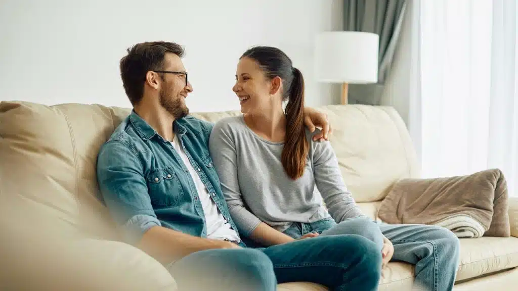 A couple sits on a couch smiling and looking into each other’s eyes.