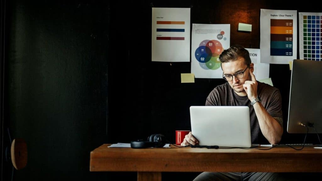 A thoughtful man in glasses looks at a laptop while sitting at a desk with papers on the wall behind him.