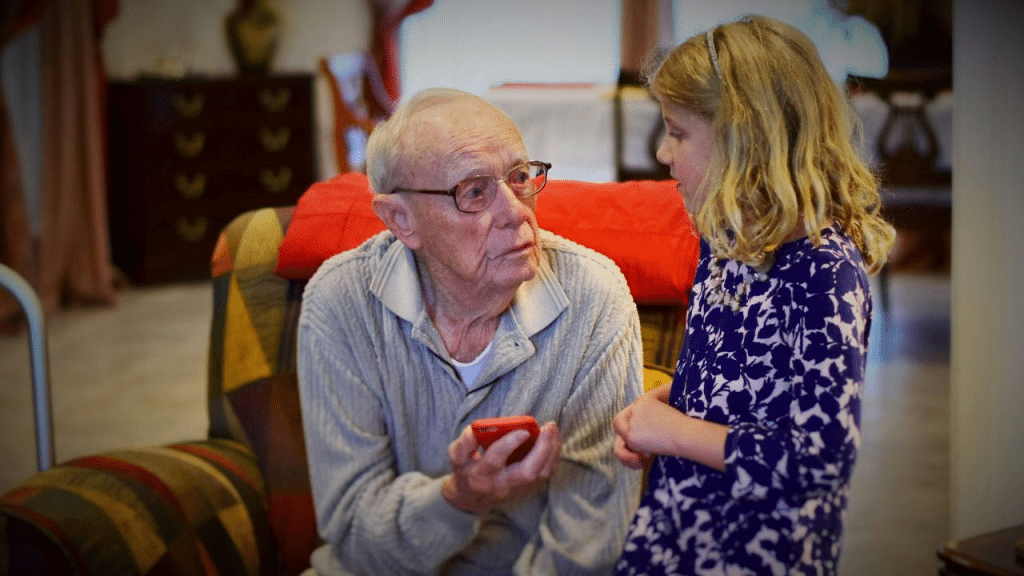 An older man in a chair holds a phone while a young girl looks at him.