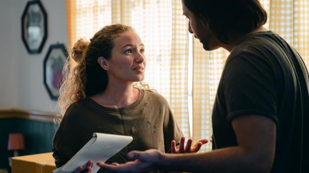 A woman with curly hair holds a notepad and talks to a man with long hair.