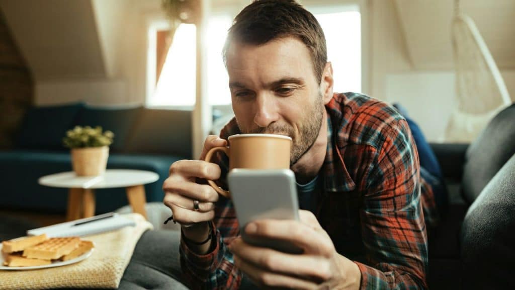A man lies on a couch, holding a coffee mug and looking at his phone.