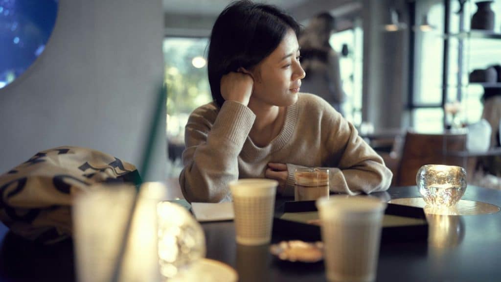 A woman with dark hair sits at a table in a cafe, looking away from the camera.
