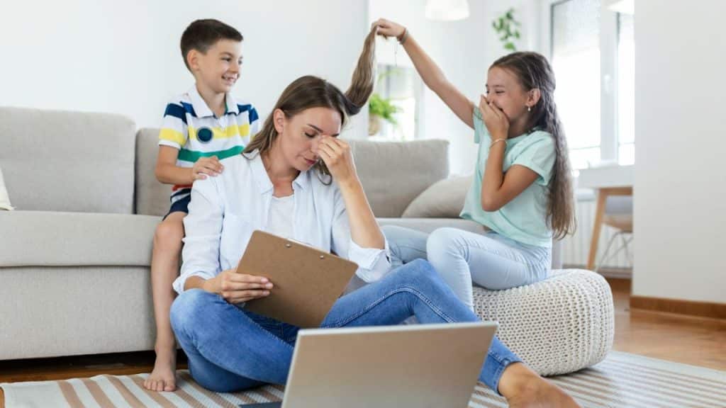 A frustrated mother sits with a laptop while two children play and pull her hair.