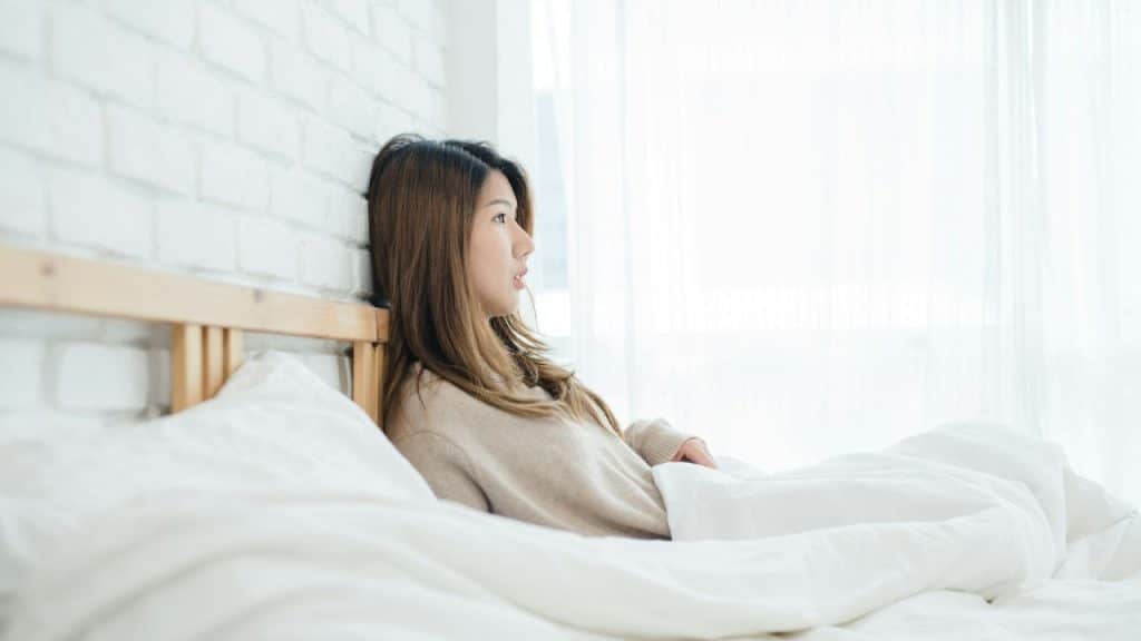 A woman with long hair sits up in bed, propped against a wooden headboard.