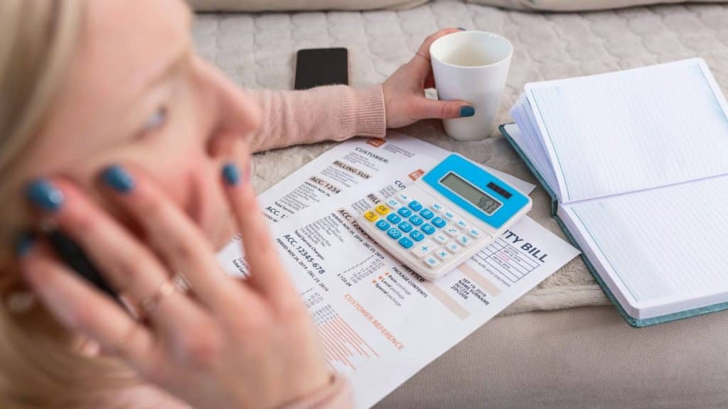 A woman holds a coffee cup and a calculator while looking at utility bills.